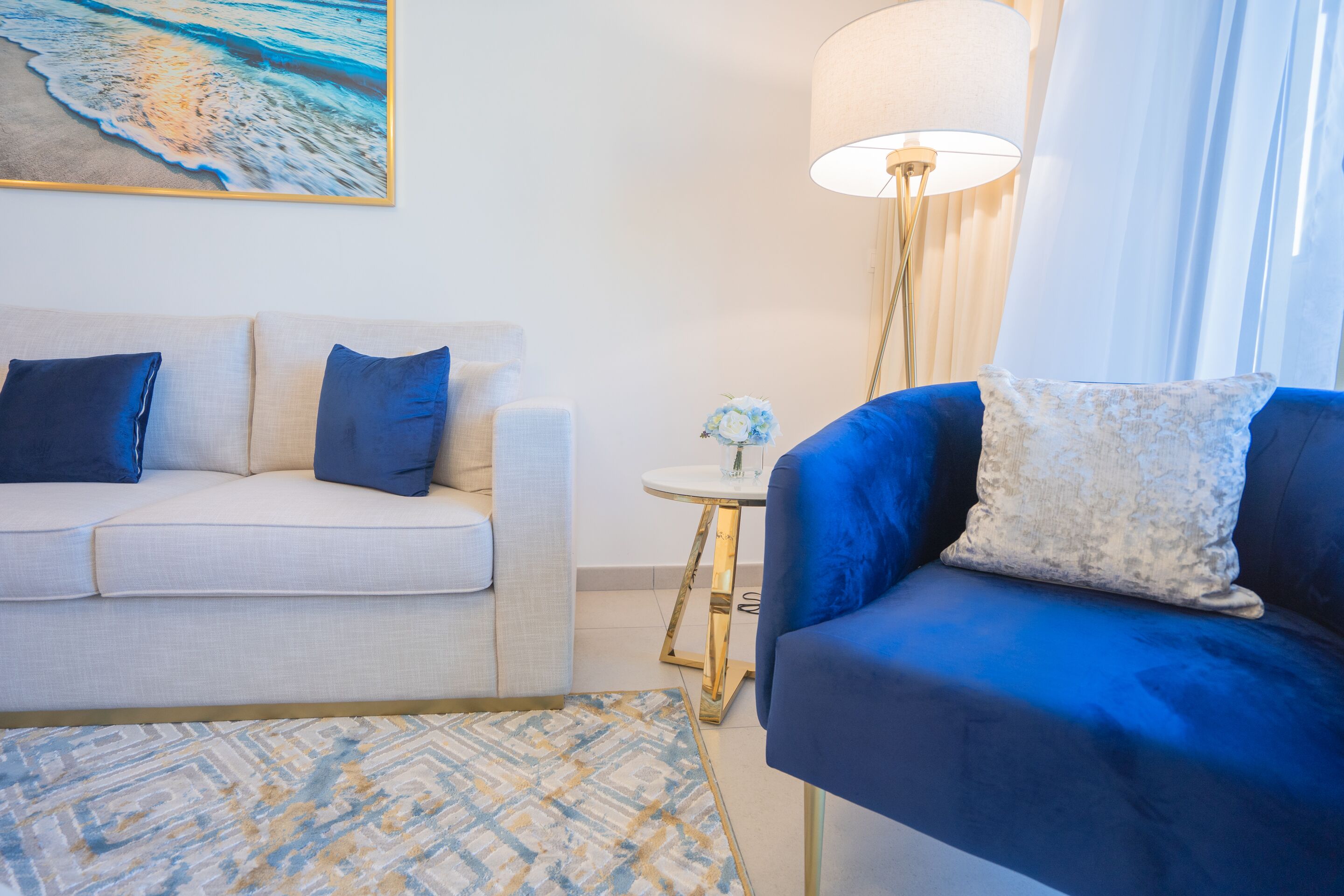 Un salon avec un canapé beige, un fauteuil bleu, un tapis à motifs, une petite table ronde avec une lampe et un tableau représentant une scène de plage.
