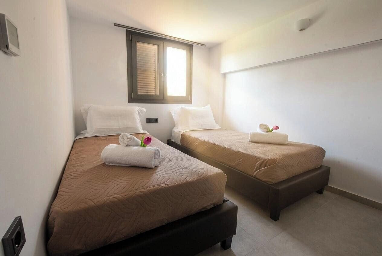 Ein Hotelzimmer mit zwei Betten, jeweils mit einer braunen Bettdecke und weißen Kissen. Es gibt ein Fenster mit Jalousien, ein Thermostat an der Wand und eine kleine Pflanze auf einem der Betten.