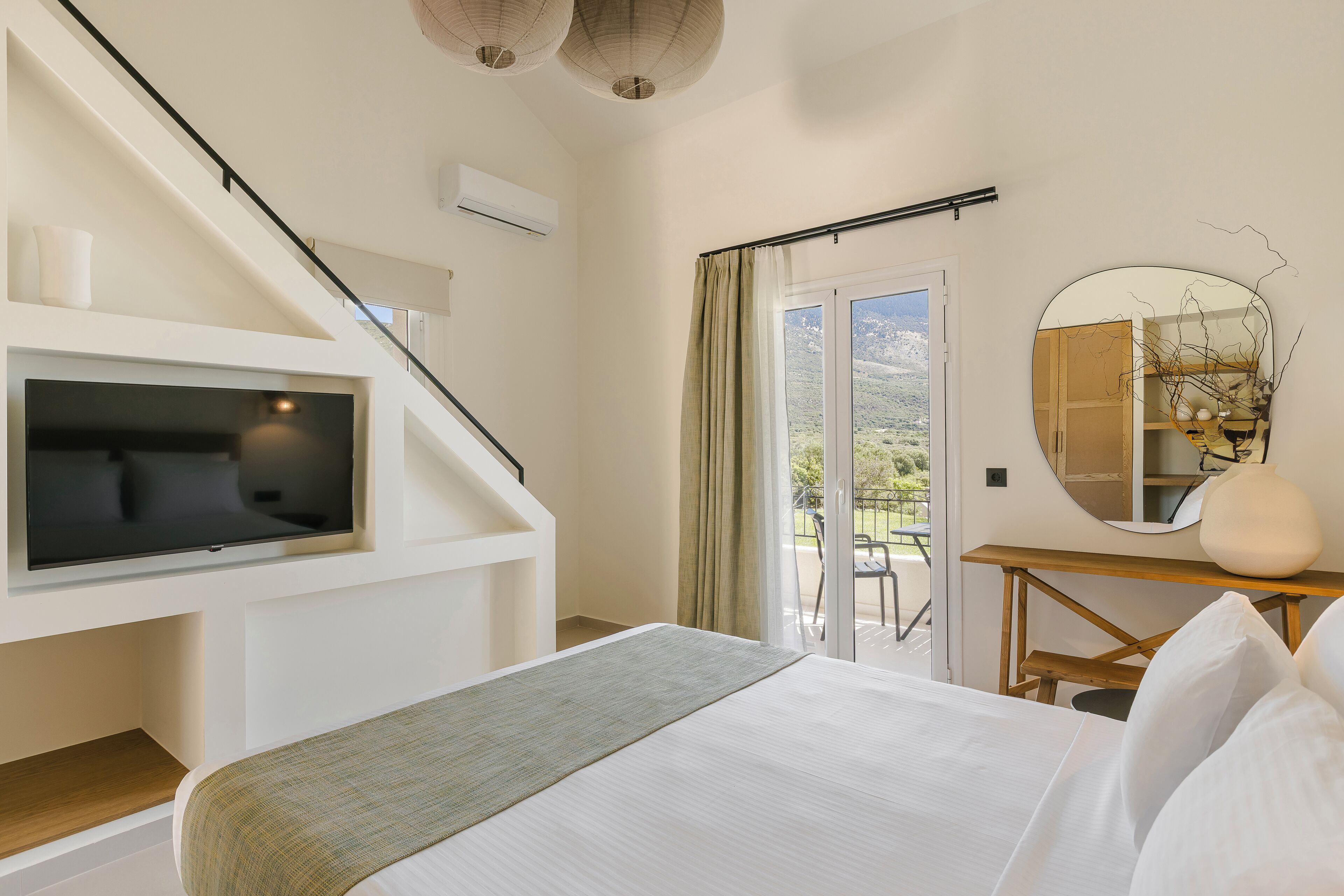 A bedroom with a bed, a TV mounted on the wall, a staircase, a balcony with a view, and a mirror on a wooden table.