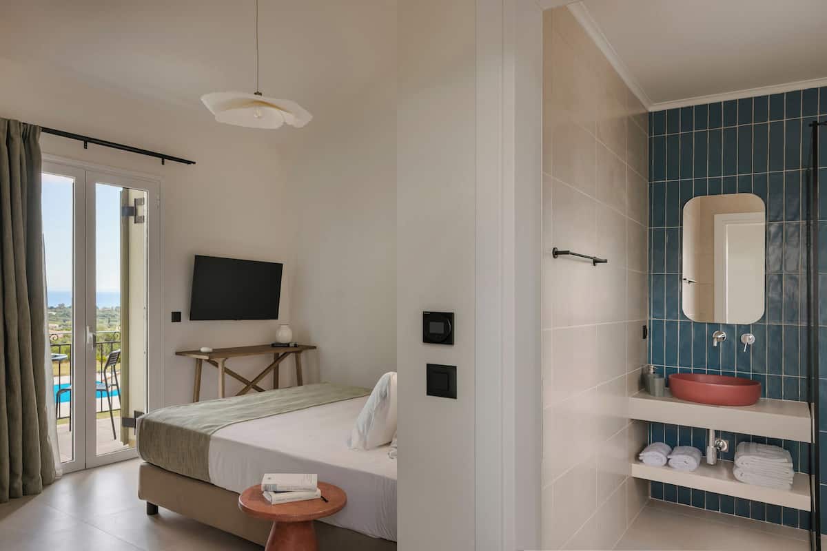 Quarto de hotel com cama, televisão, mesinha, banheiro com pia e espelho, e vista para o exterior.