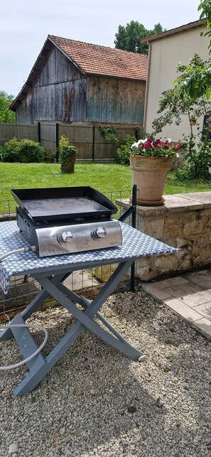 Ein Grill auf einer Tischplatte mit Metallgestell, ein Holzgebäude im Hintergrund und ein Blumentopf.