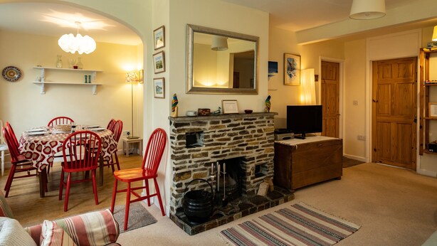A cozy living room with a stone fireplace, a dining area with red chairs, and a television on a wooden cabinet. - Pencreek (Wadebridge)