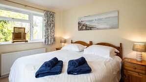 A neatly made bed with two rolled towels, a wooden headboard, a bedside lamp, and a painting of a coastal scene. - Pencreek (Wadebridge)