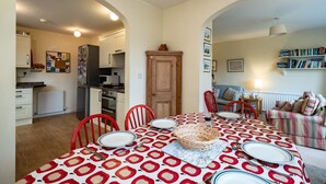 A dining area with a patterned tablecloth, red chairs, and a basket. There is a kitchen in the background with white cabinets and a refrigerator. - Pencreek (Wadebridge)