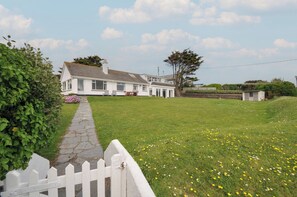A white house with a stone pathway leading to the entrance, surrounded by a grassy lawn with yellow flowers. - Dolphins (Wadebridge)