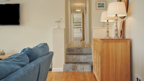Un salon avec un canapé bleu, un meuble en bois sur lequel sont posées deux lampes, un téléviseur fixé au mur et un escalier menant à une autre pièce.