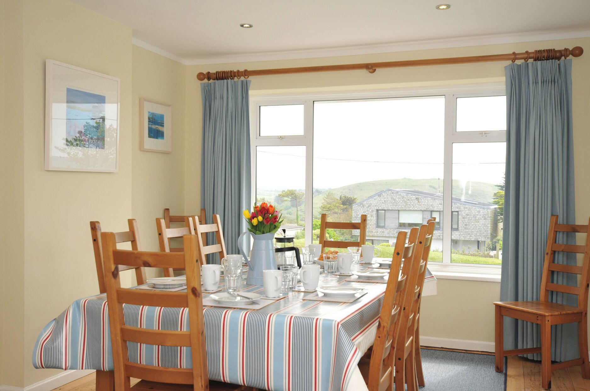 A dining room with a table set for six, wooden chairs, a vase of flowers, and a view of a house through large windows.