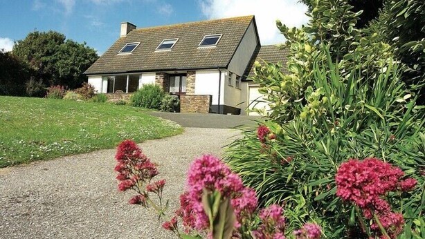 A two-story house with a sloped roof, a gravel driveway, and a garden with pink flowers. - Rosevallen (Wadebridge)