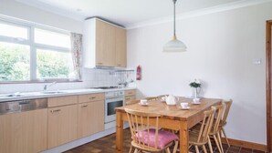 A kitchen with wooden cabinets, a dining table with chairs, a window with curtains, and a hanging light fixture. - Rosevallen (Wadebridge)