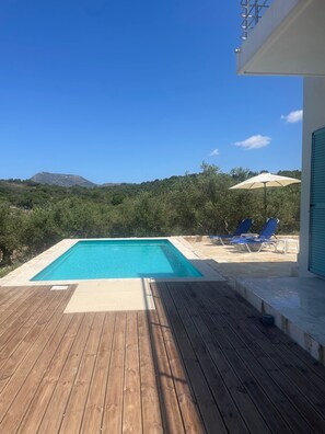 A rectangular swimming pool with a wooden deck in the foreground, a clear blue sky, and a view of green hills in the background.