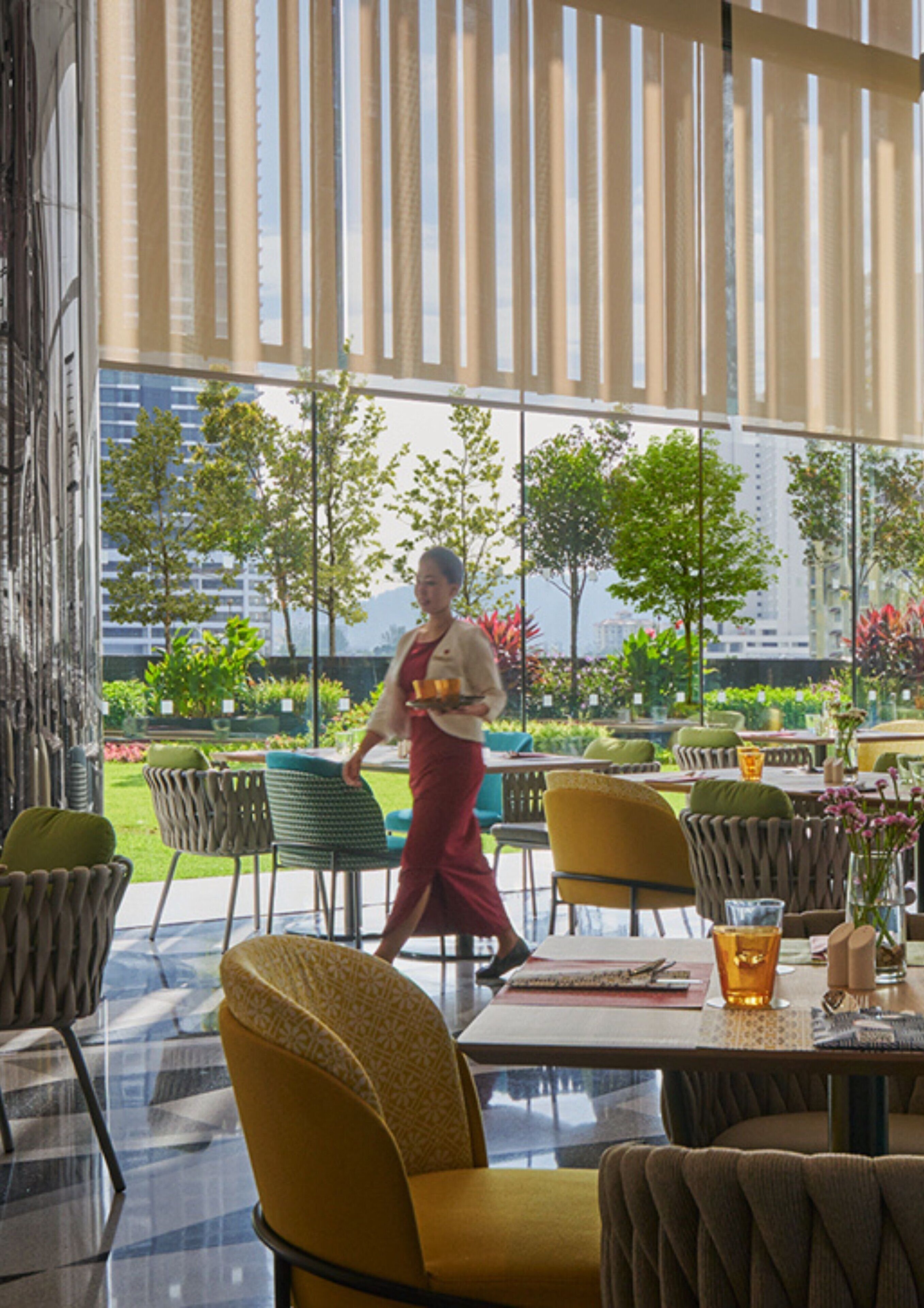 A modern restaurant with outdoor seating, large windows, and a view of greenery and buildings.