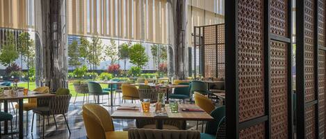 An indoor dining area with tables and chairs, large windows, and a view of greenery outside.