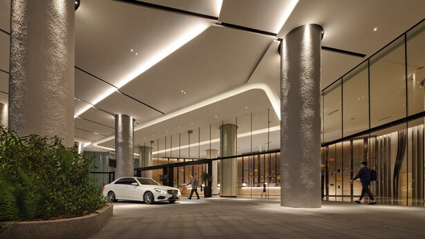 Een modern gebouw met een entree, grote pilaren, glazen deuren en een witte auto die er geparkeerd staat.