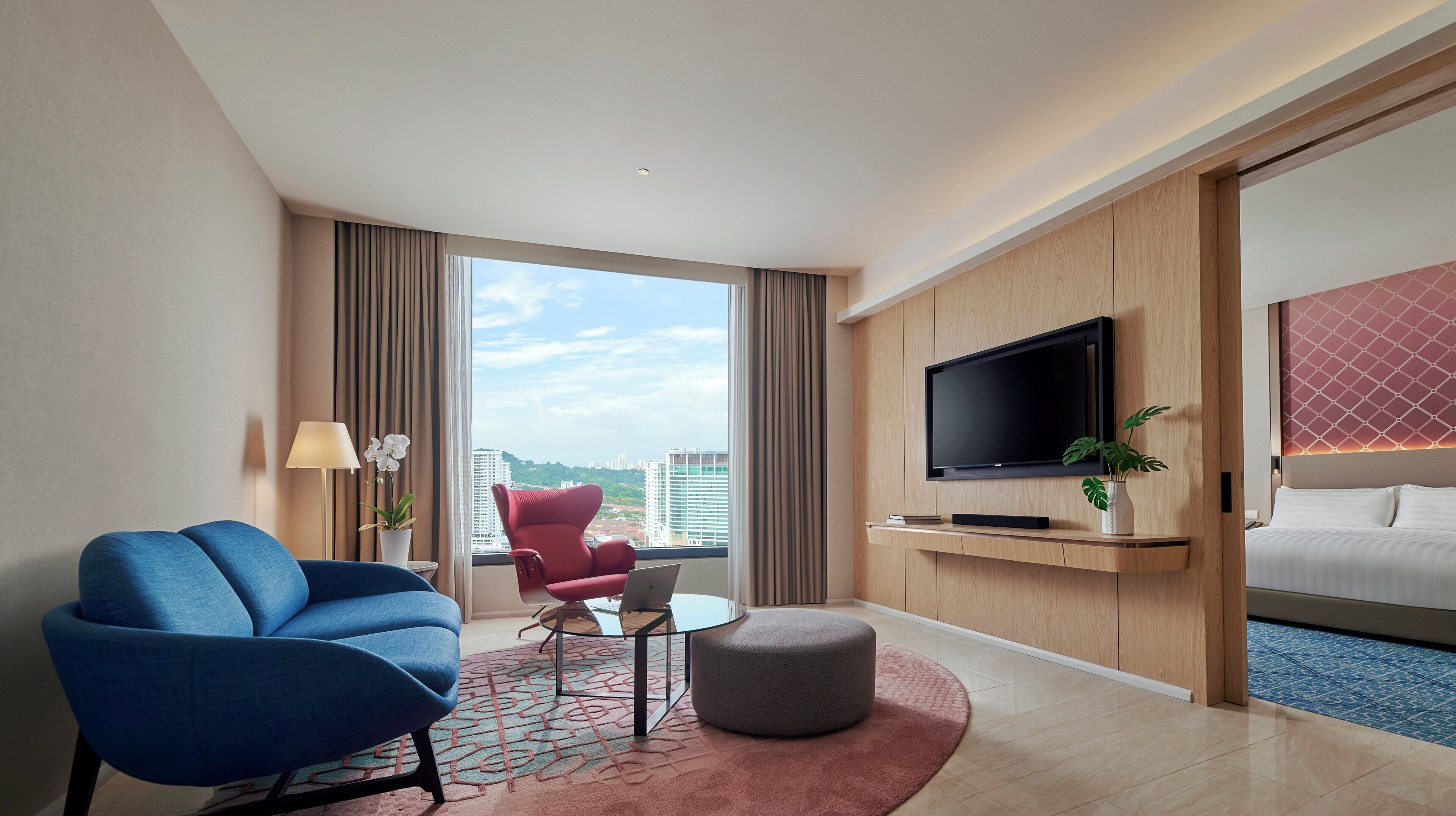 A modern hotel room with a large window, a flat-screen TV, a red chair, a blue sofa, and a bed.