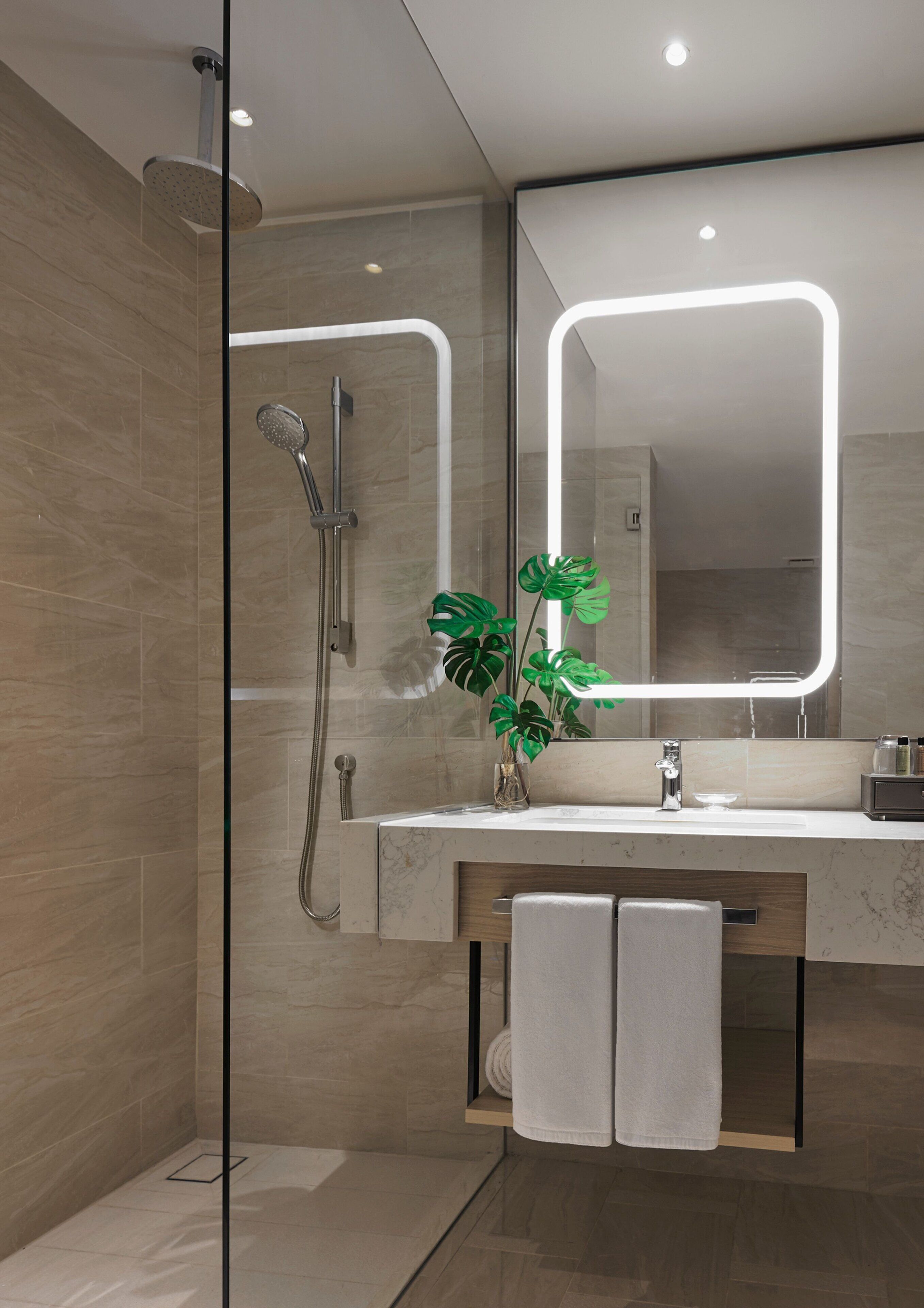 A modern bathroom with a glass-enclosed shower, a large mirror with integrated lighting, a marble countertop, and a potted plant.