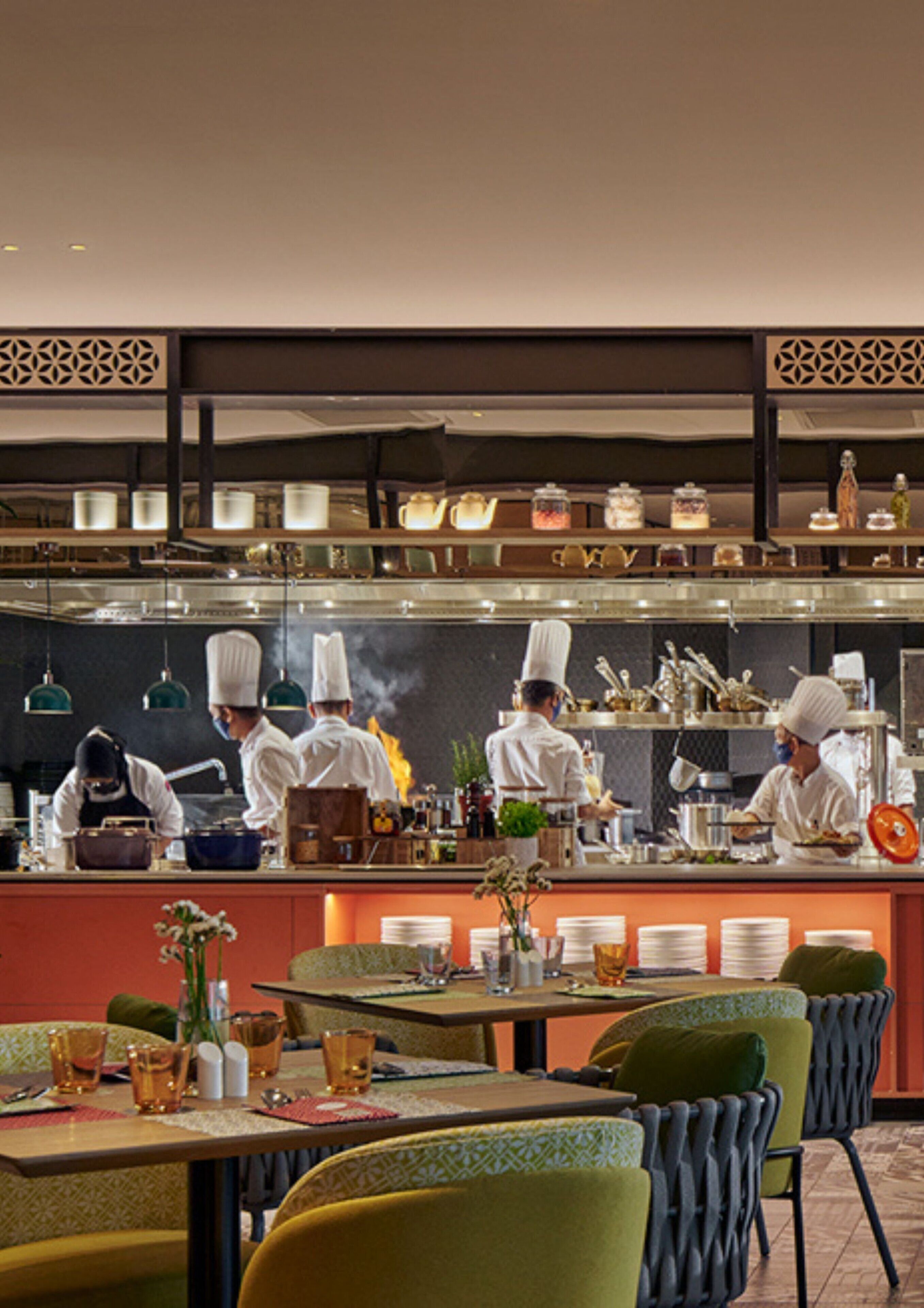 Un restaurante con zona de bar donde cocineros preparan comida. Las mesas están dispuestas con platos, vasos y cubiertos.