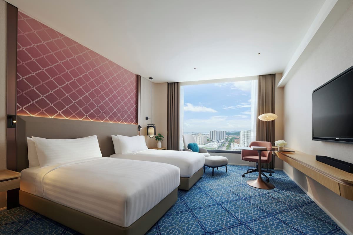 A hotel room with two beds, a desk, a TV, and a large window with a city view.