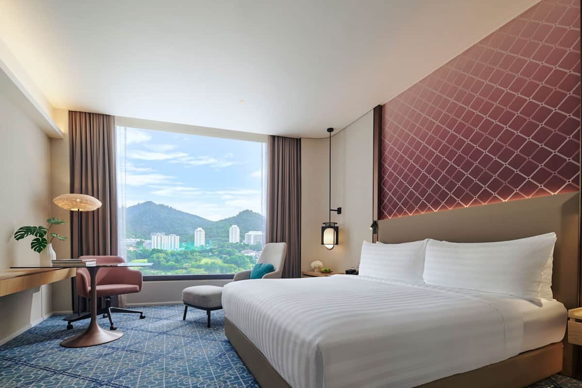 A hotel room with a large bed, a desk with a chair, a small table, a lamp, and a view of buildings and mountains through the window.