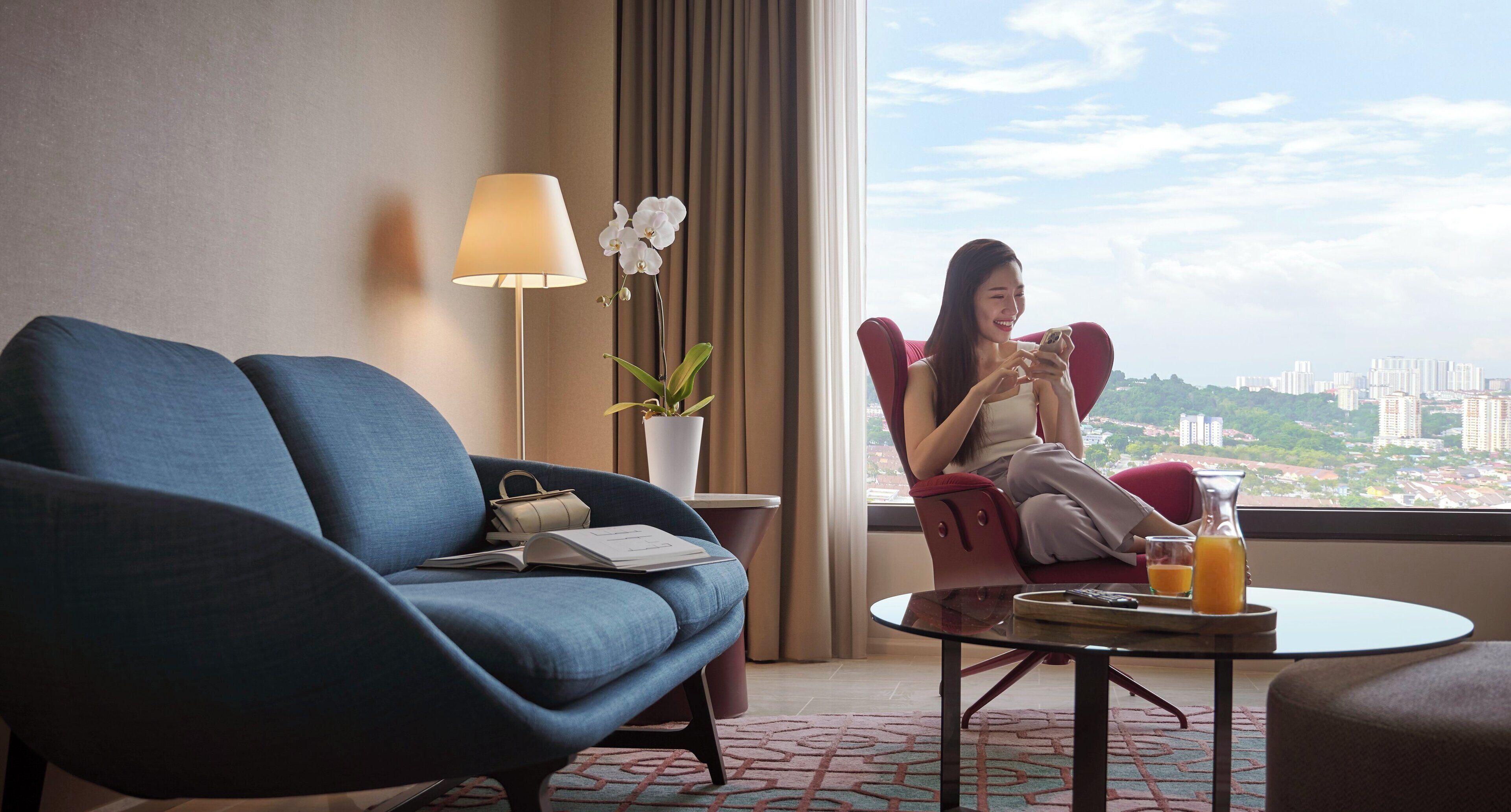 Quarto de hotel moderno com sofá azul, poltrona vermelha, pequena mesa com um copo de suco de laranja e vista da cidade através de grandes janelas.
