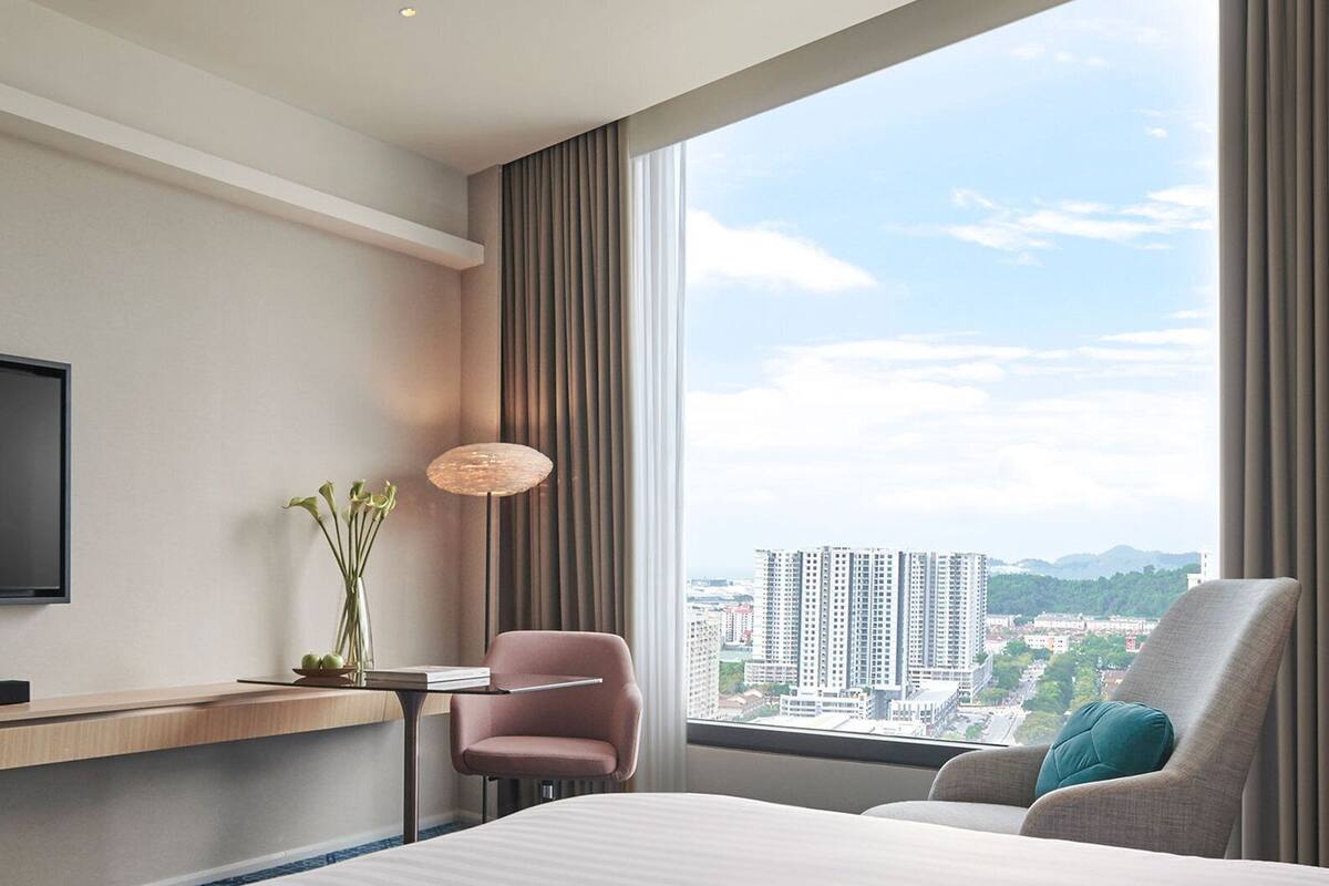 Hotellihuone, jossa on suuri ikkuna, sänky, työpöytä, tuoli ja televisio.