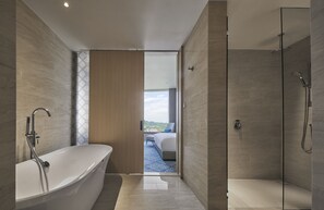 A modern bathroom with a freestanding bathtub, a glass-enclosed shower, and a view of a bedroom with a bed and a window.
