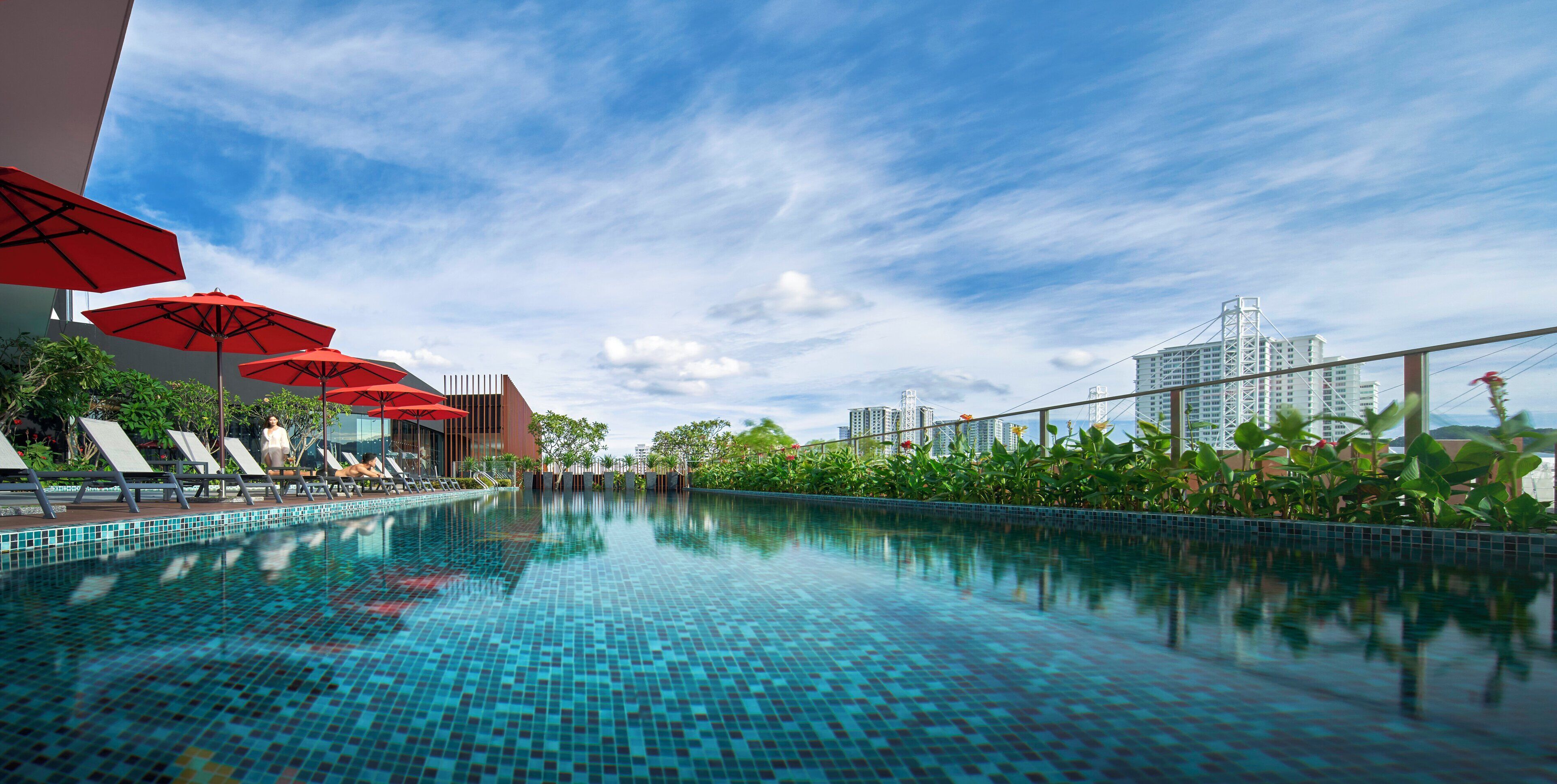 A swimming pool with tiled flooring, red umbrellas, and lounge chairs.