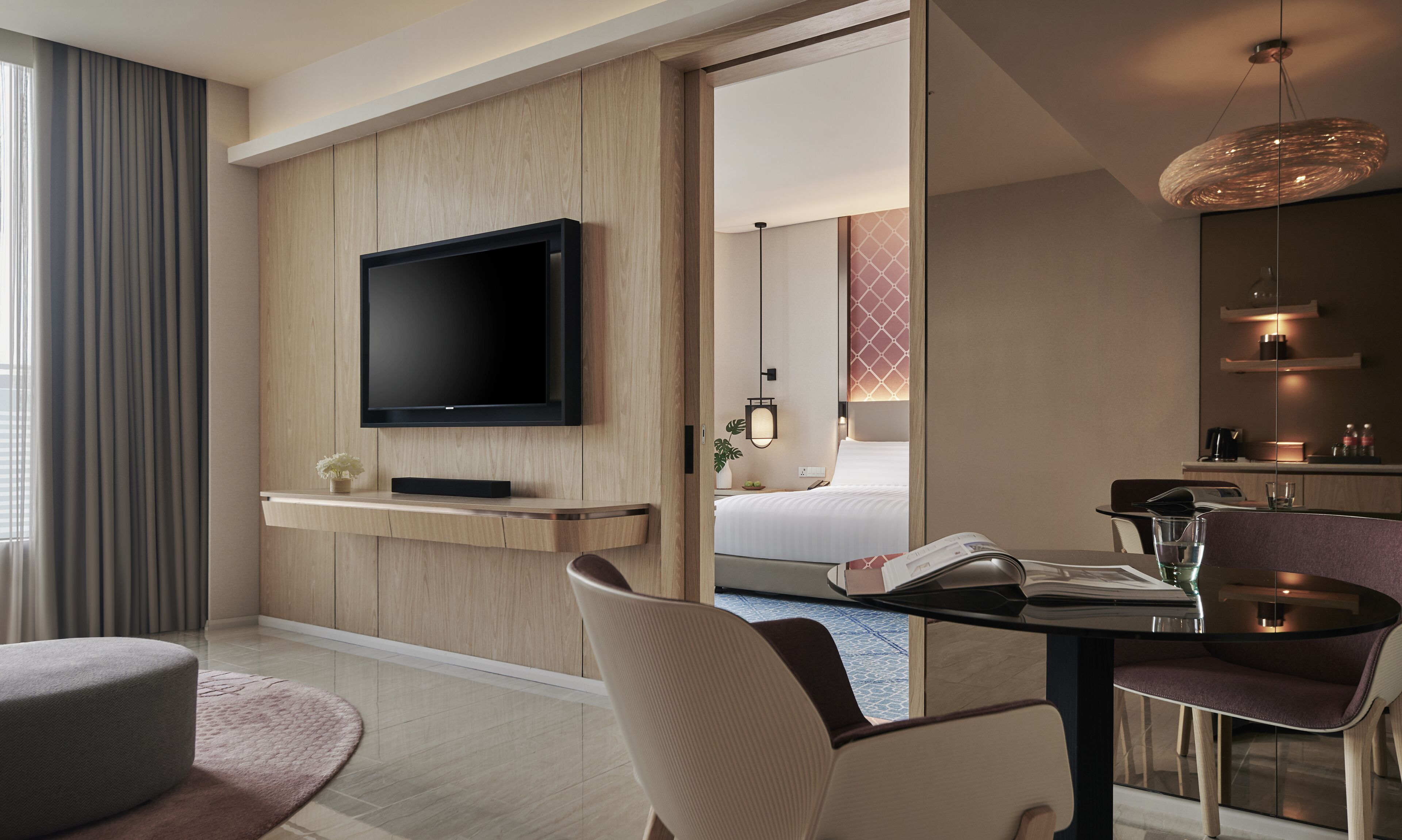 Een moderne hotelkamer met een flatscreen televisie bevestigd aan een houten paneel, een eettafel met stoelen, een klein zithoekje met een beklede bank, en een zichtbare badkamer met een douche.