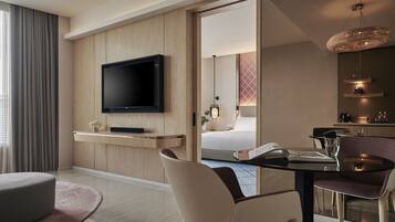 Een moderne hotelkamer met een flatscreen televisie bevestigd aan een houten paneel, een eettafel met stoelen, een klein zithoekje met een beklede bank, en een zichtbare badkamer met een douche.