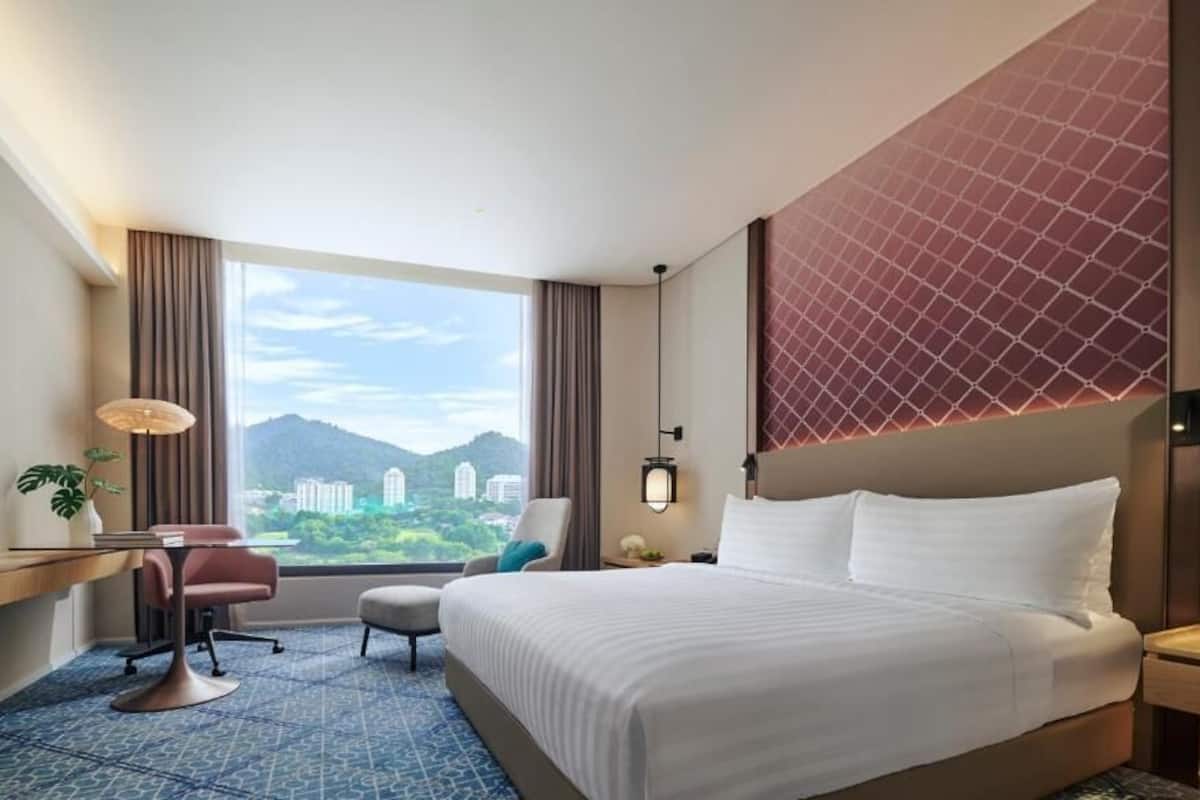 A hotel room with a large bed, a desk with a chair, a small table, and a view of mountains through the window.