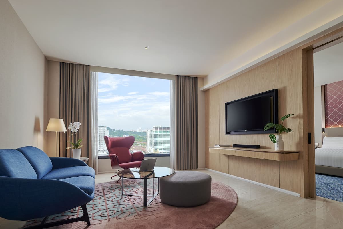 A modern hotel room with a large window, a flat-screen TV, a blue sofa, a red armchair, and a bed with a patterned headboard.