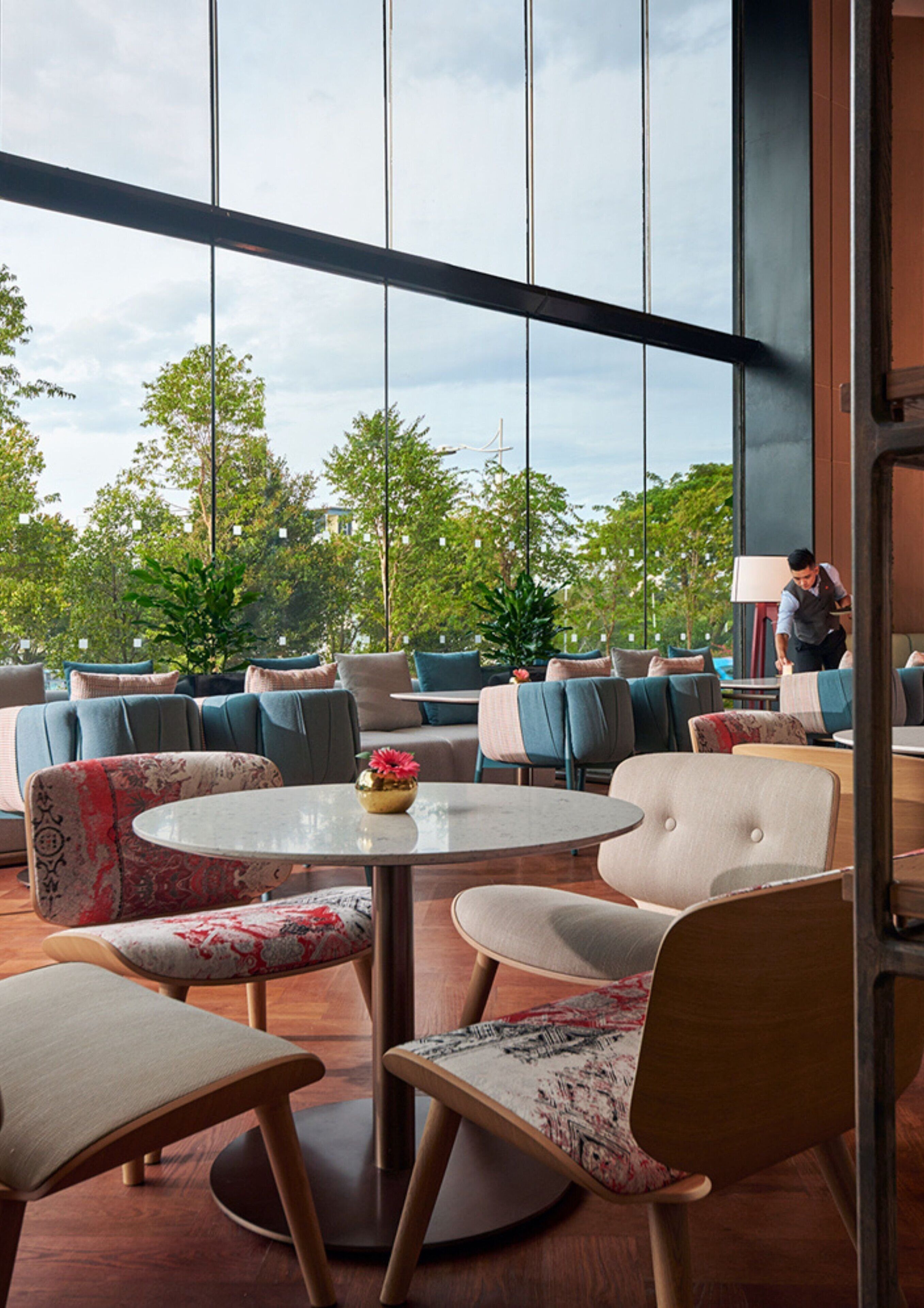 A modern lounge area with round tables and comfortable seating, large windows offering a view of greenery and a building in the distance.