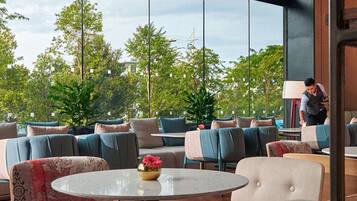 A modern lounge area with round tables and comfortable seating, large windows offering a view of greenery and a building in the distance.