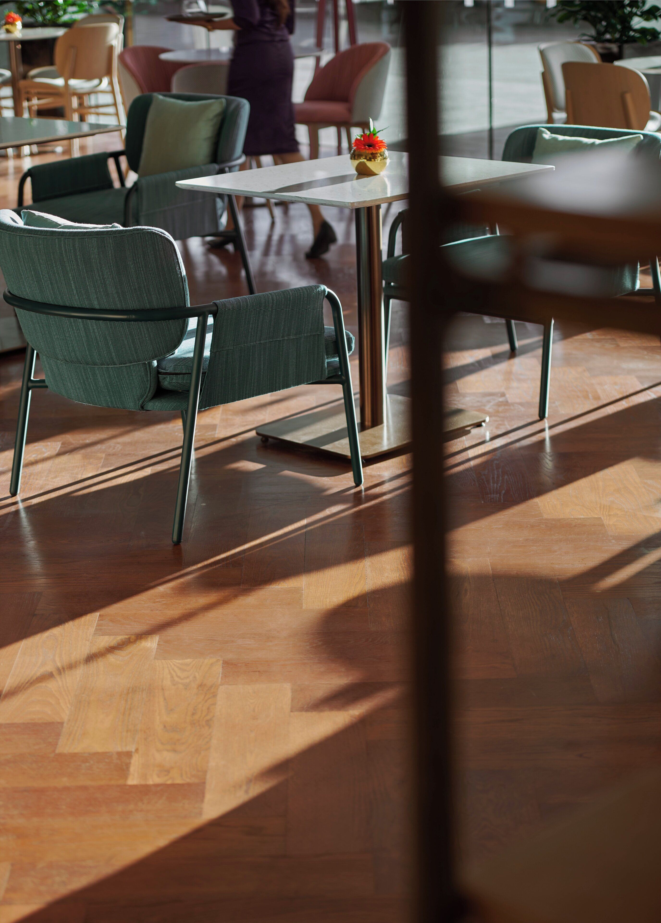 A modern indoor seating area with herringbone wooden flooring, green upholstered chairs, and a small table with a flower centerpiece.