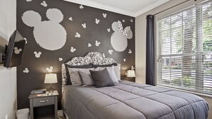 Une chambre à coucher avec un lit, des tables de chevet, un autocollant mural à l’effigie de Mickey Mouse et une fenêtre avec des stores.