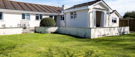 Una casa de dos pisos con fachada blanca y techo marrón. Un pequeño jardín delantero con seto y patio empedrado.