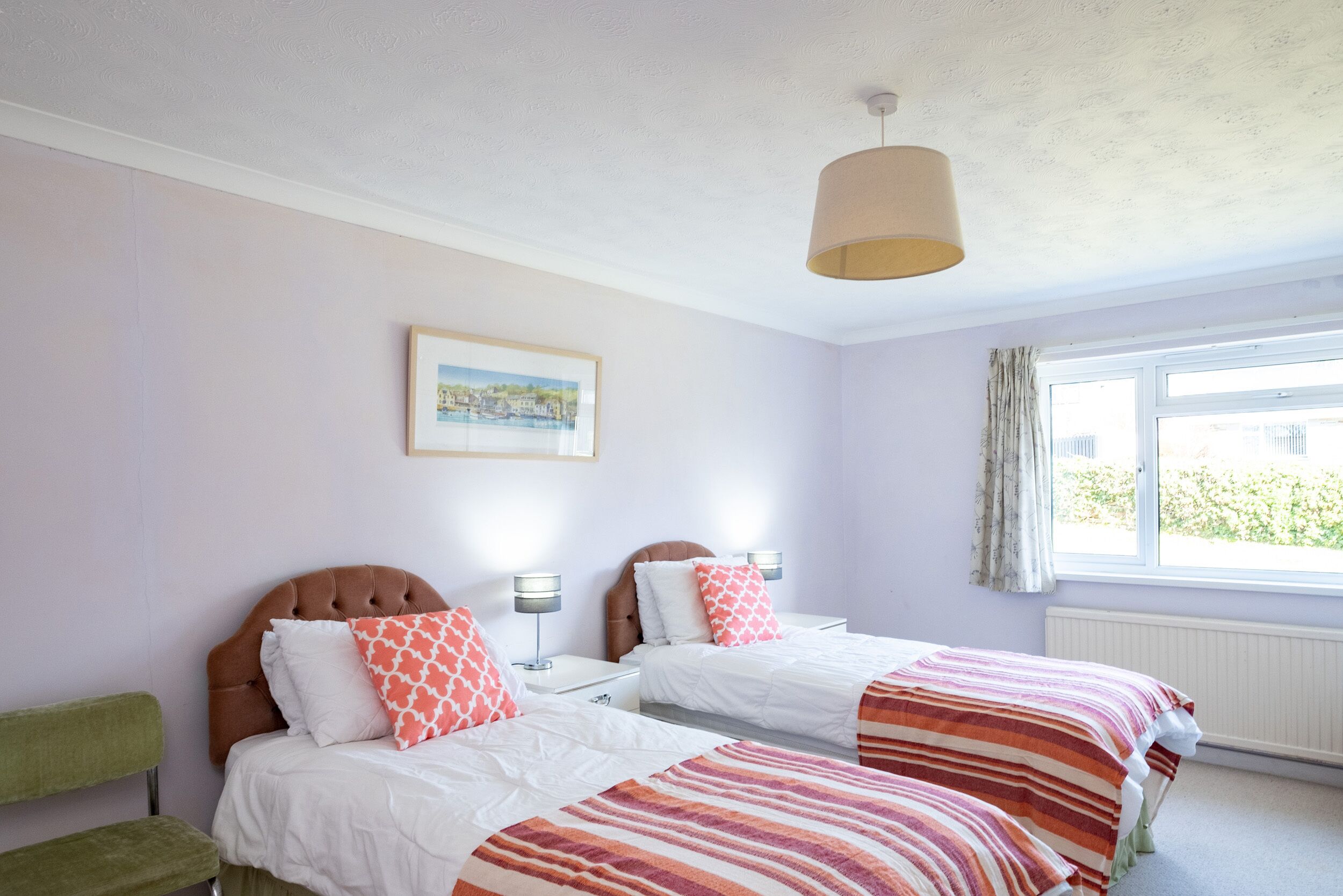 Une chambre avec deux lits, un fauteuil vert, une lampe suspendue, une fenêtre avec des rideaux et un tableau encadré au mur.