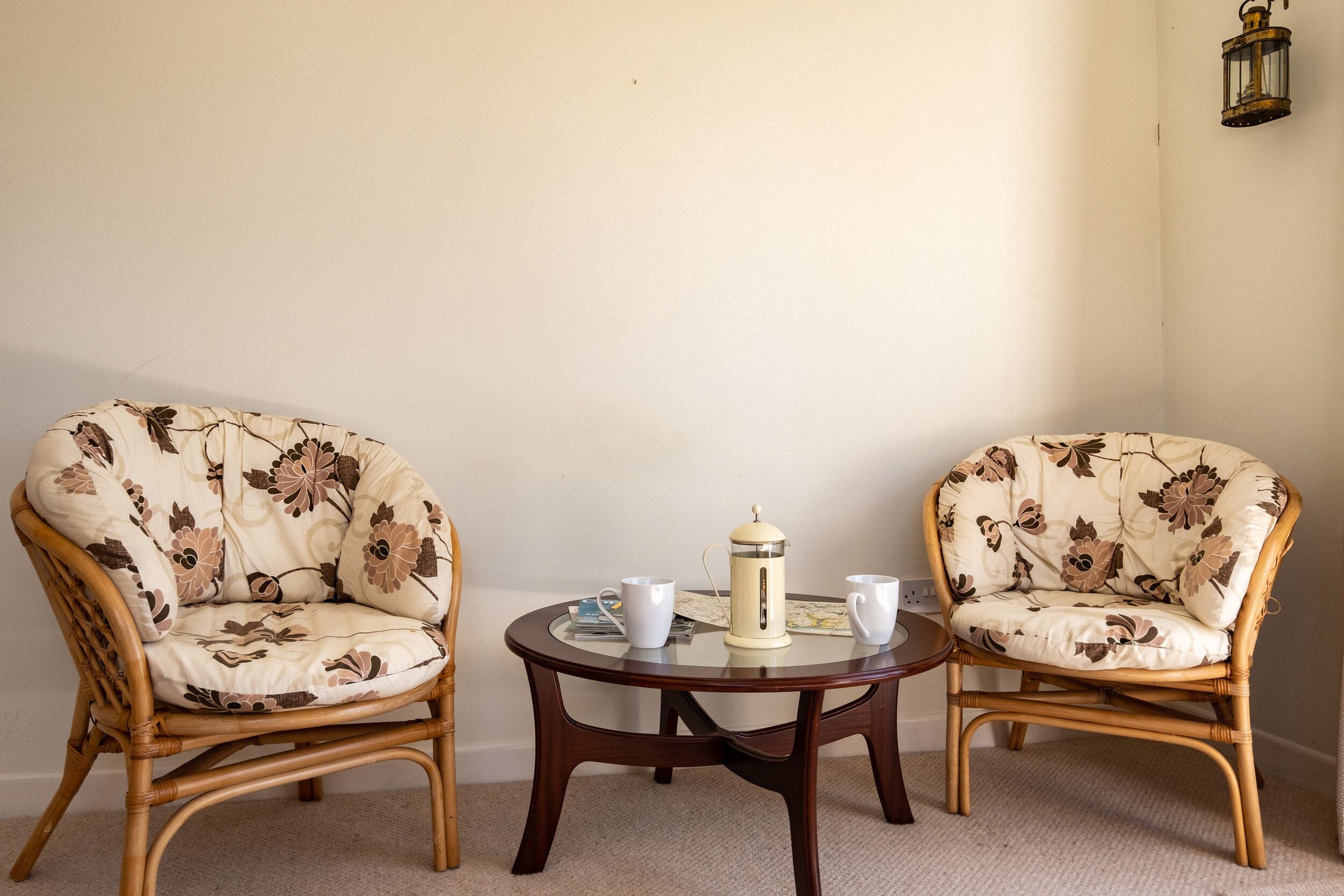 Two wicker chairs with floral upholstery, a small round wooden table with a coffee pot and two white cups, and a wall-mounted lantern.