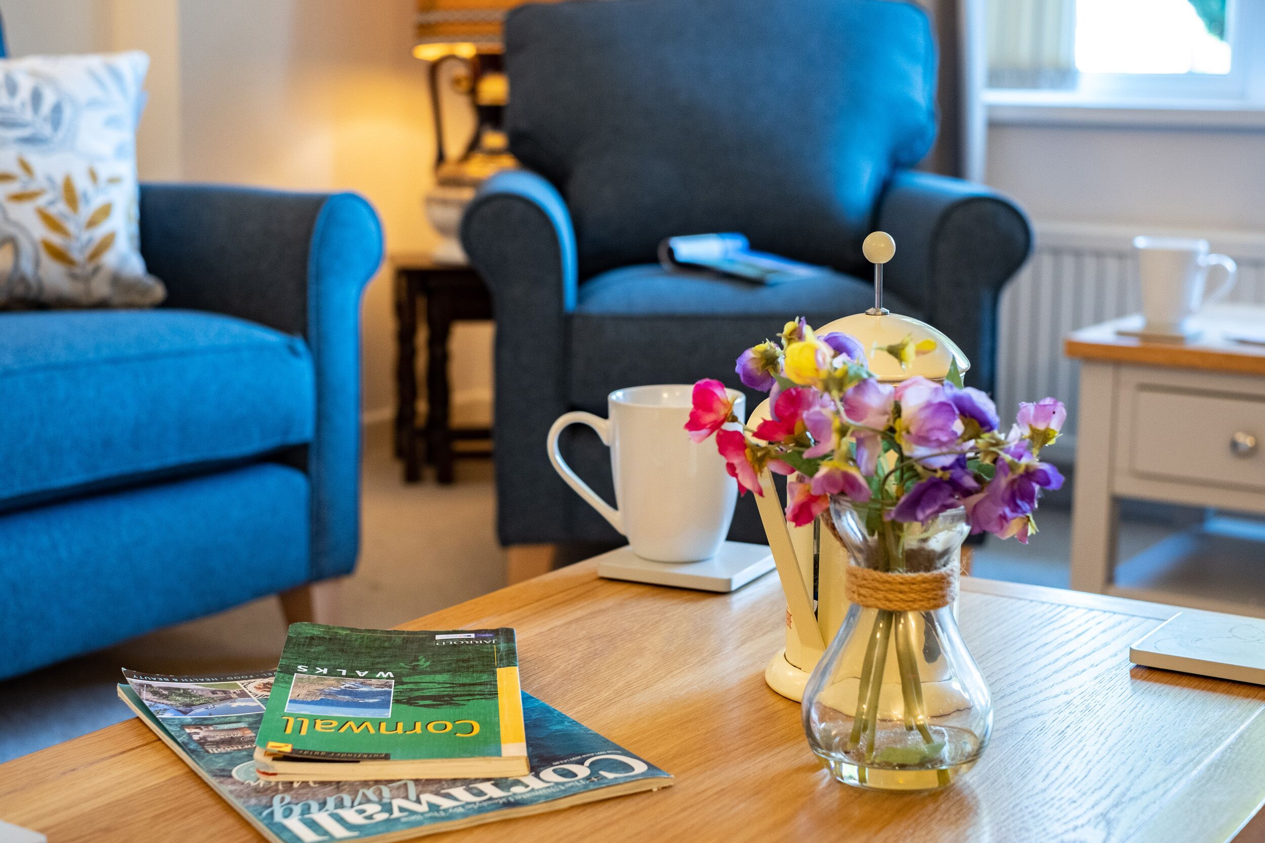 A living room with a blue sofa, a coffee table with a magazine, a white mug, and a vase with flowers.