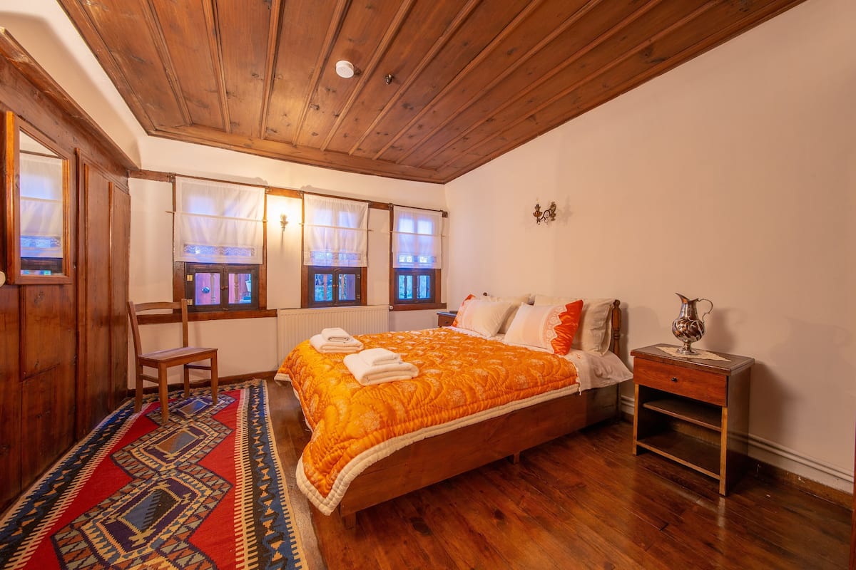 A bedroom with a wooden ceiling, a bed with an orange quilt, a nightstand, and a chair.