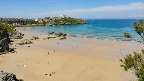 A beach with clear blue water, rocky shore, and buildings on a hill in the background. - 49 Atlantic Reach (Newquay)