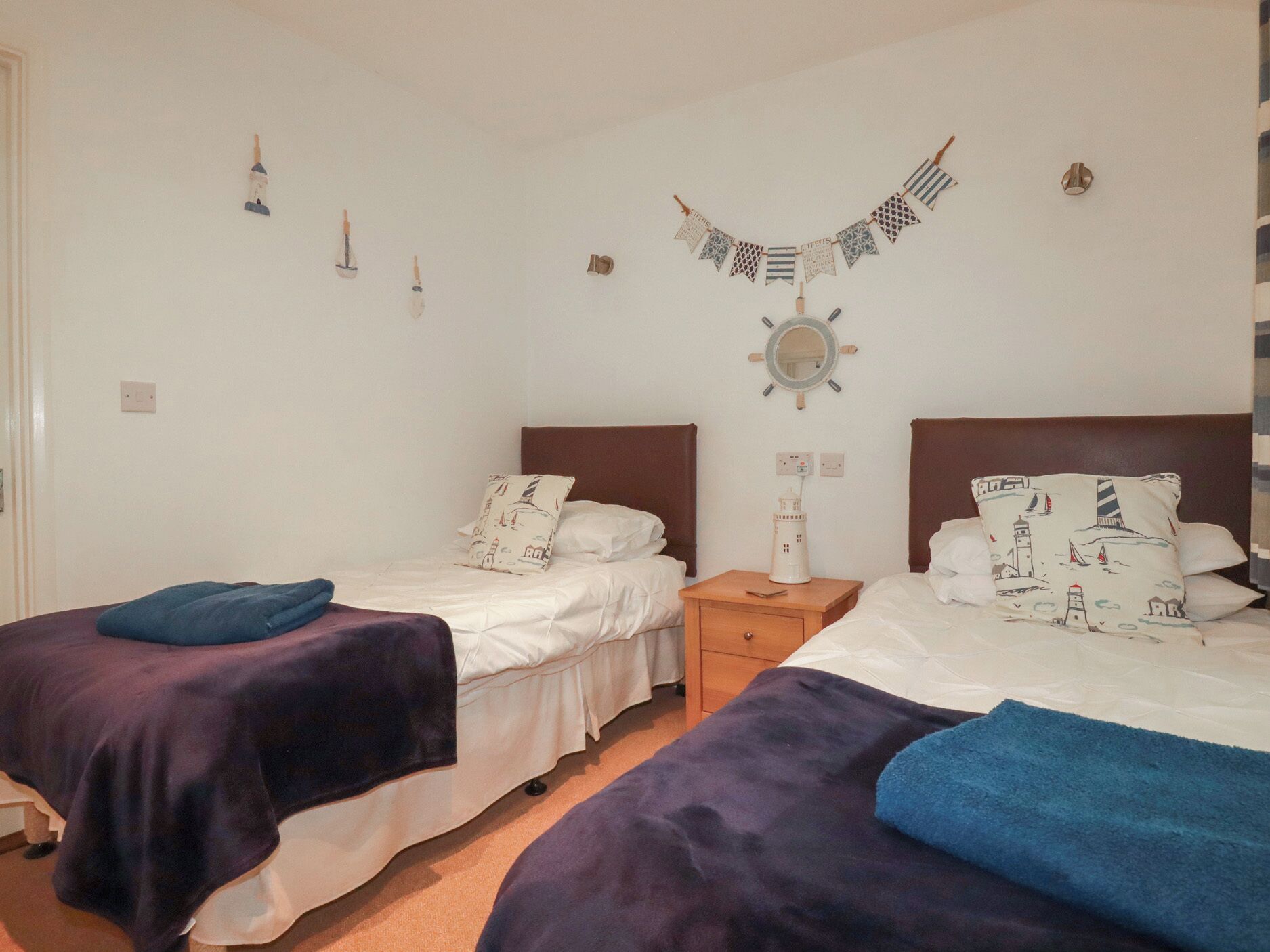 A room with two beds, each with a headboard and pillows, a bedside table with a lamp, a wall clock, and a decorative wall hanging.