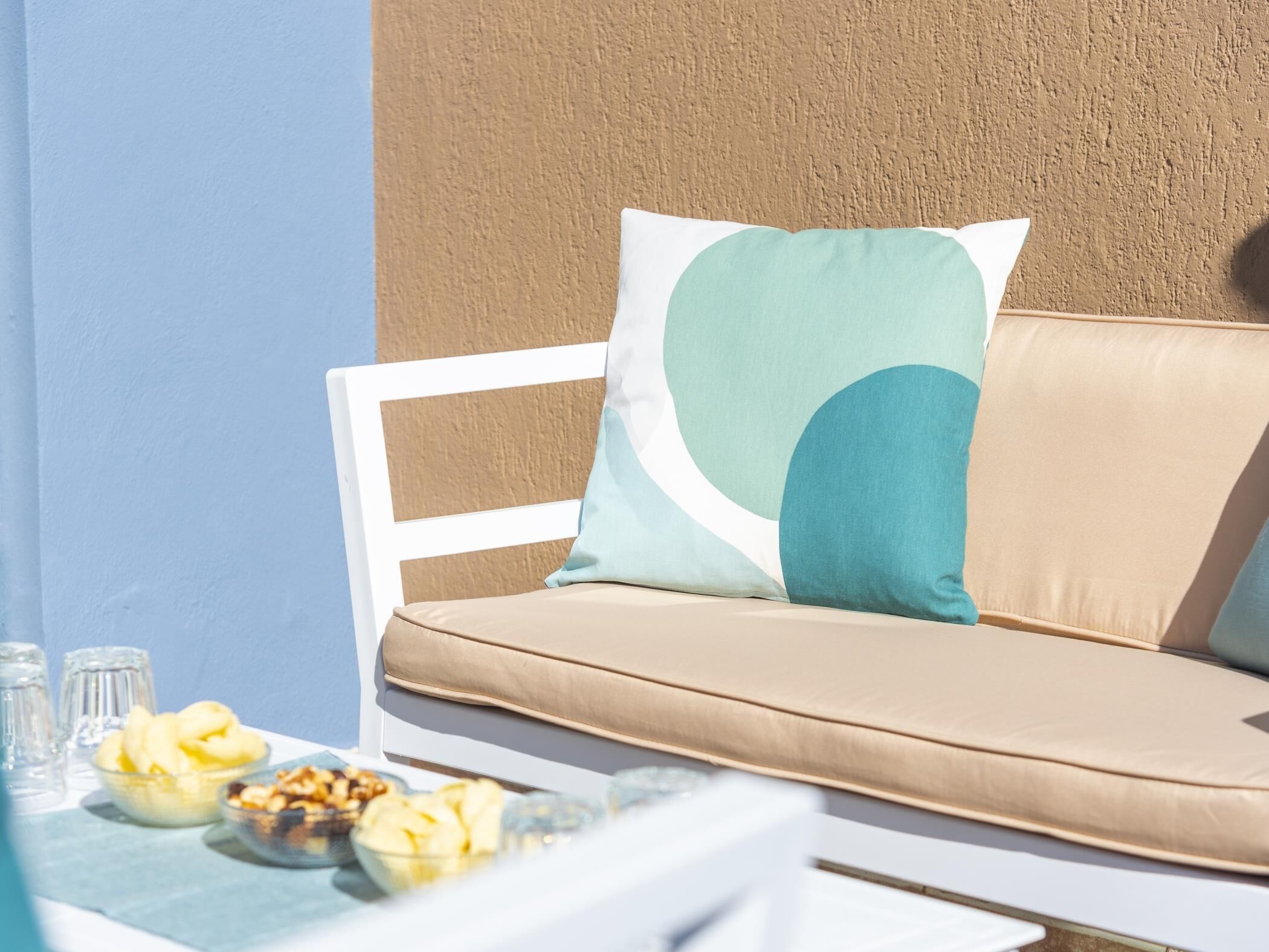 Un canapé blanc avec des coussins beige et des oreillers turquoise, une table en verre sur laquelle se trouvent des aliments, et un mur texturé en arrière-plan.