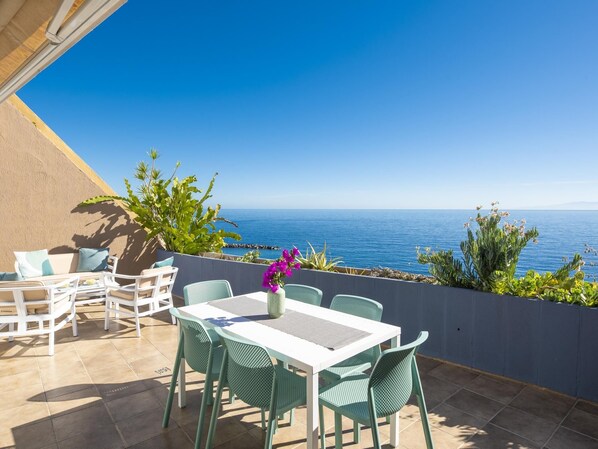 Une terrasse avec une table et des chaises, une vue sur la mer et quelques plantes.