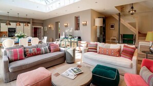 Ein geräumiges Wohnzimmer mit hoher Decke, Oberlicht und Blick zur Küche. Es gibt zwei Sofas, einen Couchtisch und einen roten Sessel.