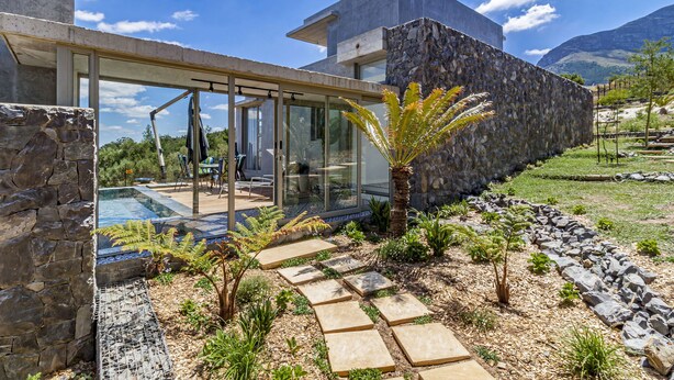 A modern house with a glass-enclosed patio, stone walls, and a landscaped garden with stepping stones and plants. - the Ultimate Venue to Relax, Breath & Recharge (Cape Town)