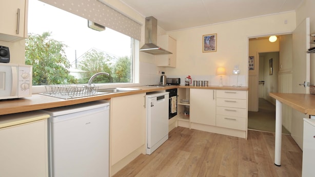 A compact kitchen with white appliances, a window with a polka dot blind, and a view of greenery outside. - Avoca (Wadebridge)