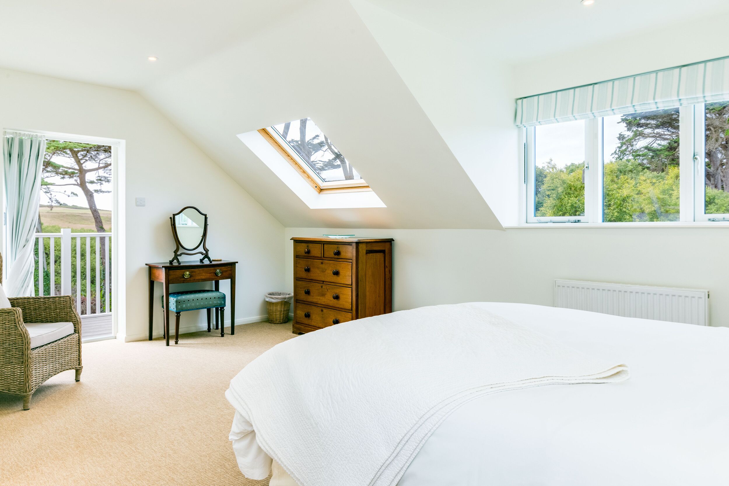 A bedroom with a skylight, a large bed, a dresser, a chair, and a window with a view of trees.