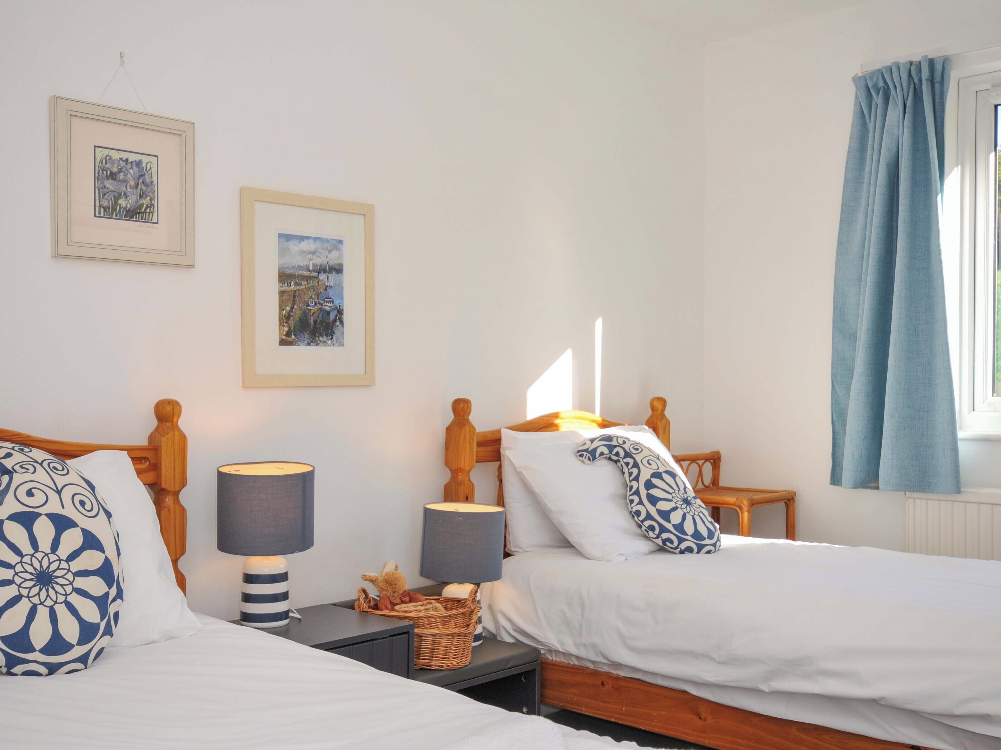 A double room with two beds, each with a wooden headboard, white bedding, and patterned pillows. There are two bedside tables with lamps and a small basket on one. Two framed pictures are hung on the wall above the beds.