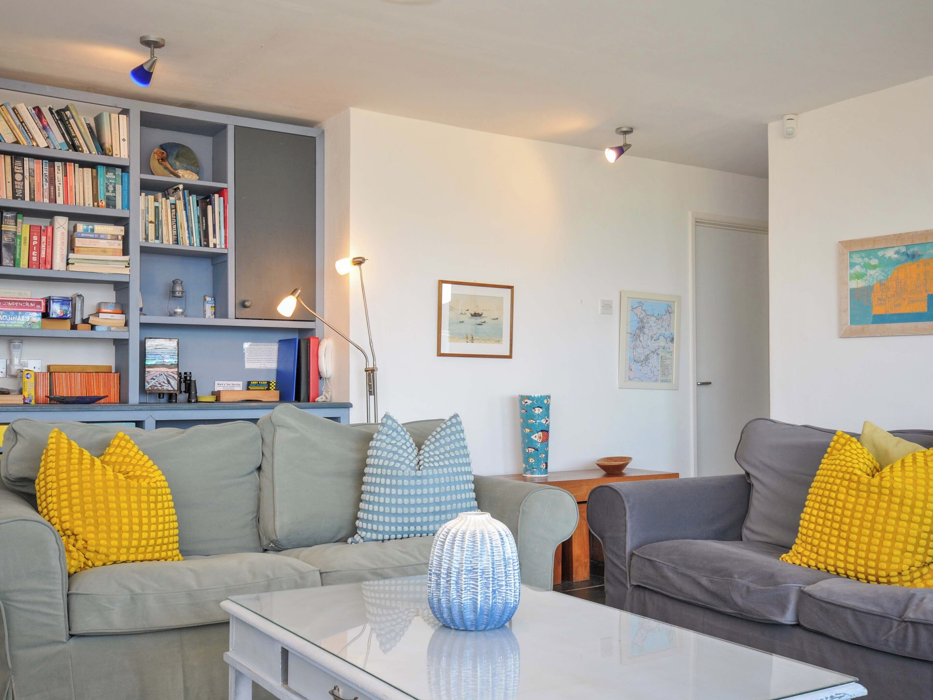 Una sala moderna con dos sofás grises, almohadas amarillas, una mesa de centro de vidrio y una estantería llena de libros y adornos.