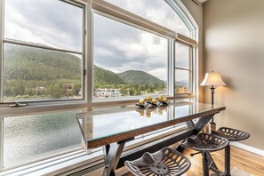 A dining area with a glass table, two stools, and a view of a lake and mountains through large windows. - Keystone Lake & Mountain Views from Private Patio & Easy Access to Activities (Keystone)